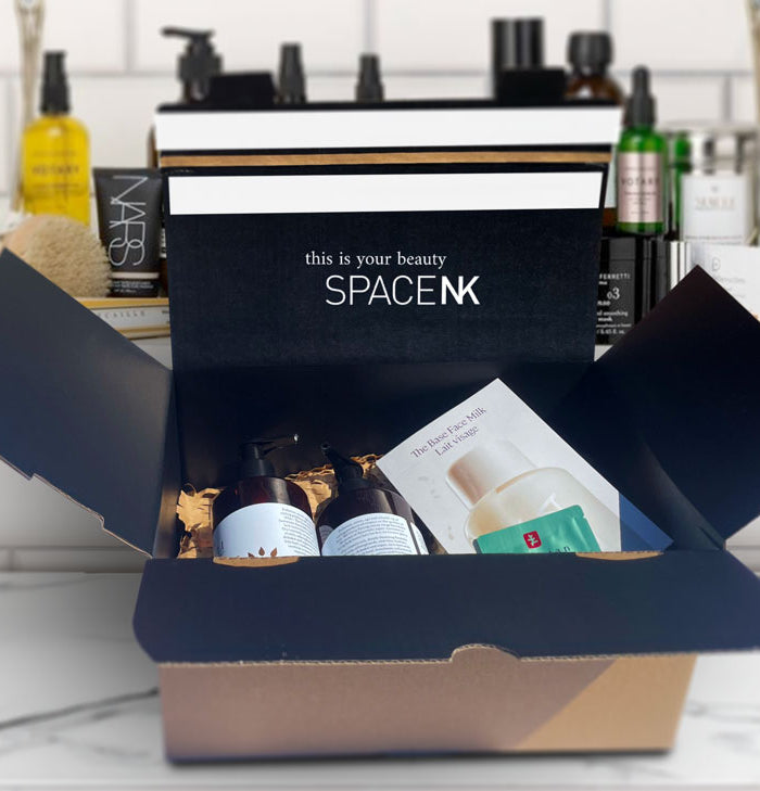 Space NK e-commerce packaging, custom printed with their logo and brand colour. The box is open showing make up products inside, sitting on top of a marble makeup counter with a shelving unit backdrop containing more cosmetic products.