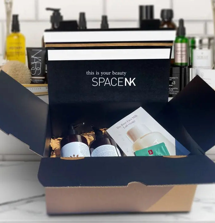 Space NK e-commerce packaging, custom printed crash lock box with their logo and brand colour. The box is open showing make up products inside, sitting on top of a marble makeup counter with a shelving unit backdrop containing more cosmetic products.