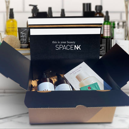 Space NK e-commerce packaging, custom printed with their logo and brand colour. The box is open showing make up products inside, sitting on top of a marble makeup counter with a shelving unit backdrop containing more cosmetic products.