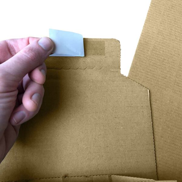 Close up of hot melt glue and peel and seal release paper being peeled back by a hand. Glue strip is on the top edge of a corrugated book wrap